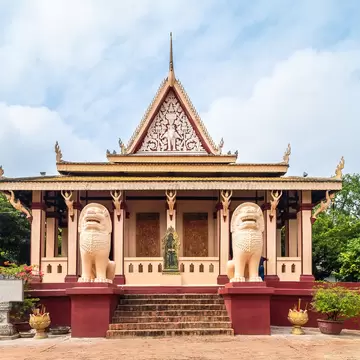 Cream and maroon Khmer temple with a tall carved gable, two white stone lions and brick steps, framed by trees