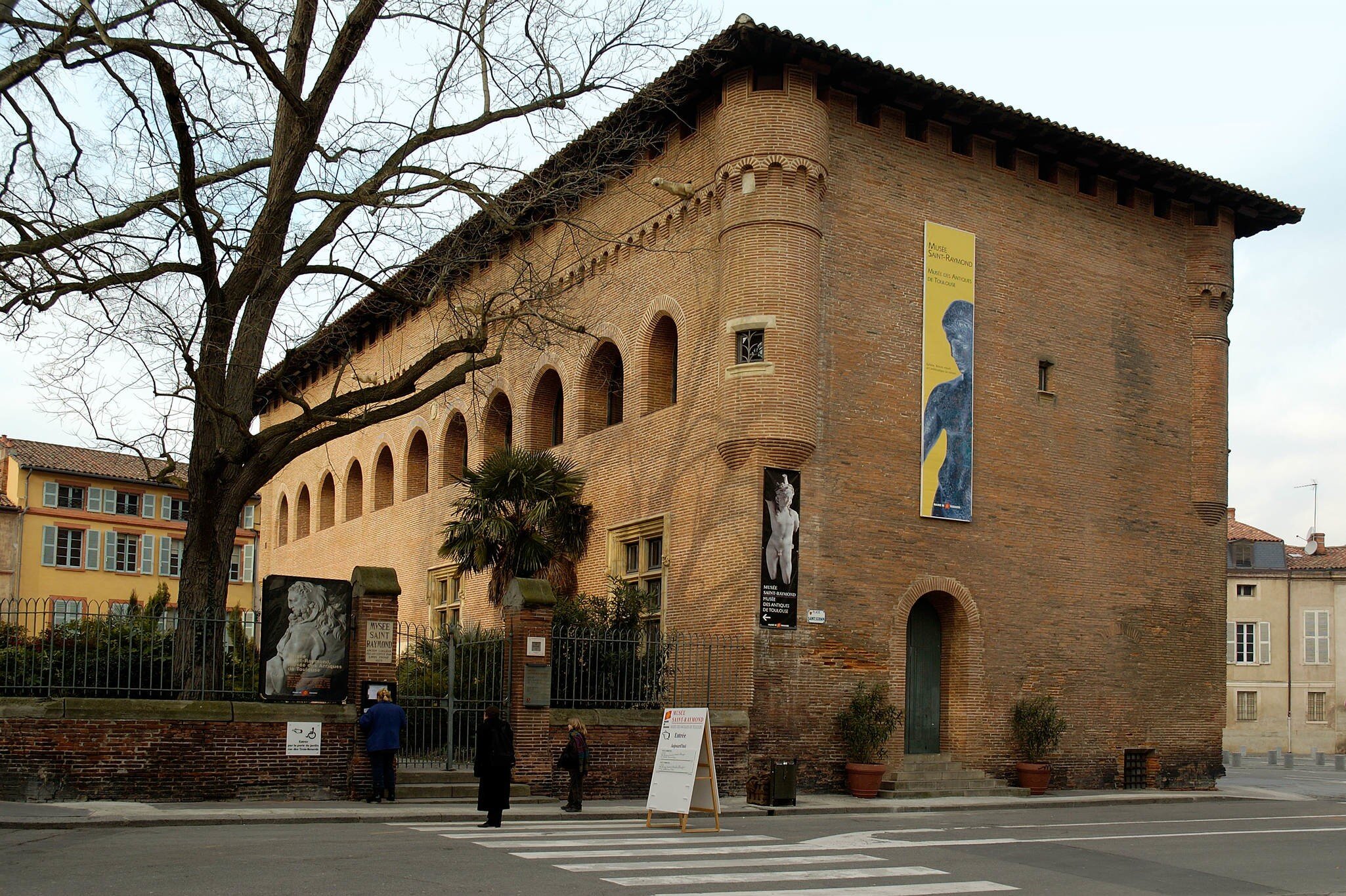 Brick medieval building with an arcaded upper floor and exhibition banners beside a city street.
