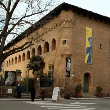 Brick medieval building with an arcaded upper floor and exhibition banners beside a city street.