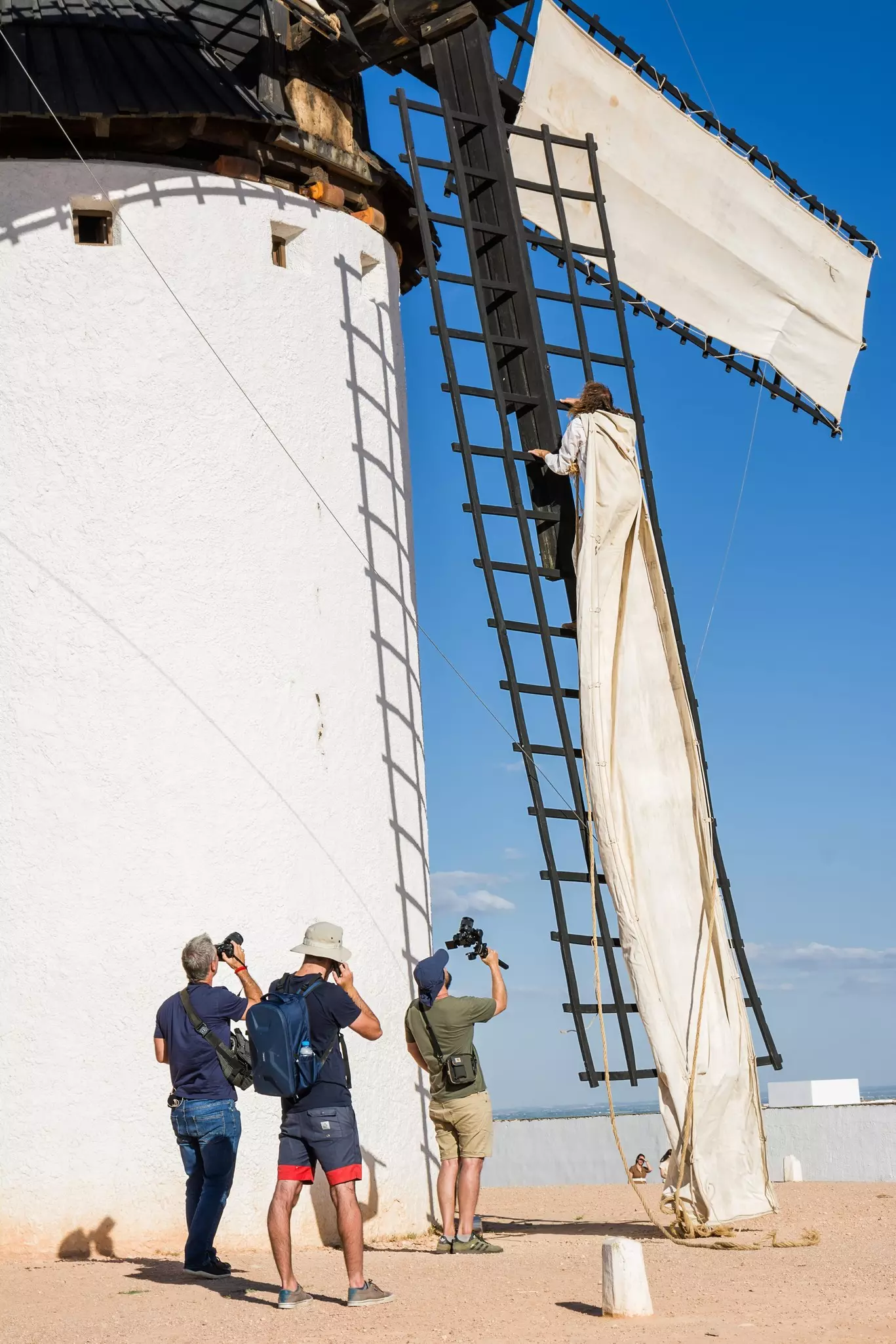 Campo de Criptana, Spain - June 22, 2022: Windmill with man climbing on the blades to install the cloth to make it work and photographers and video makers doing the shooting, License Type: media, Download Time: 2025-03-21T20:03:15.000Z, User: rhylton_redventures, Editorial: true, purchase_order: 65050 - Digital Destinations and Articles, job: Lonely Planet wip, client: Lonely Planet wip, other: Rhianydd Hylton