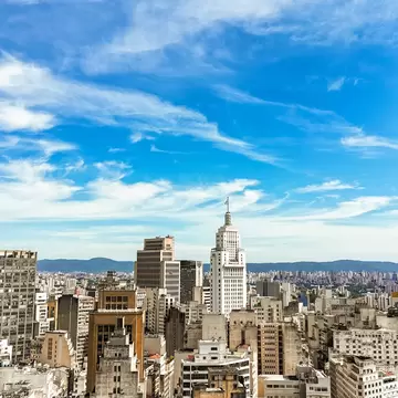 A white stepped art-deco tower standing above a dense grid of city blocks, with hills on the horizon under a streaked blue sky.