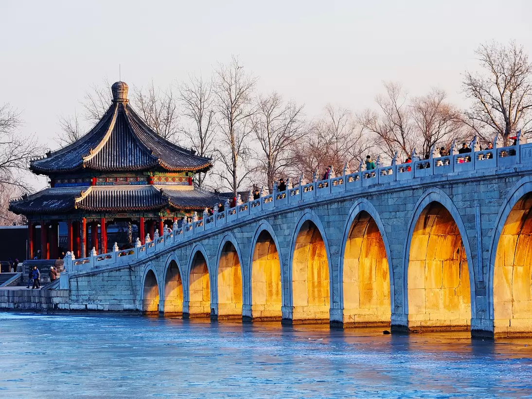 An arched bridge over water leads to a structure with a peaked roof; light shines on the archways.