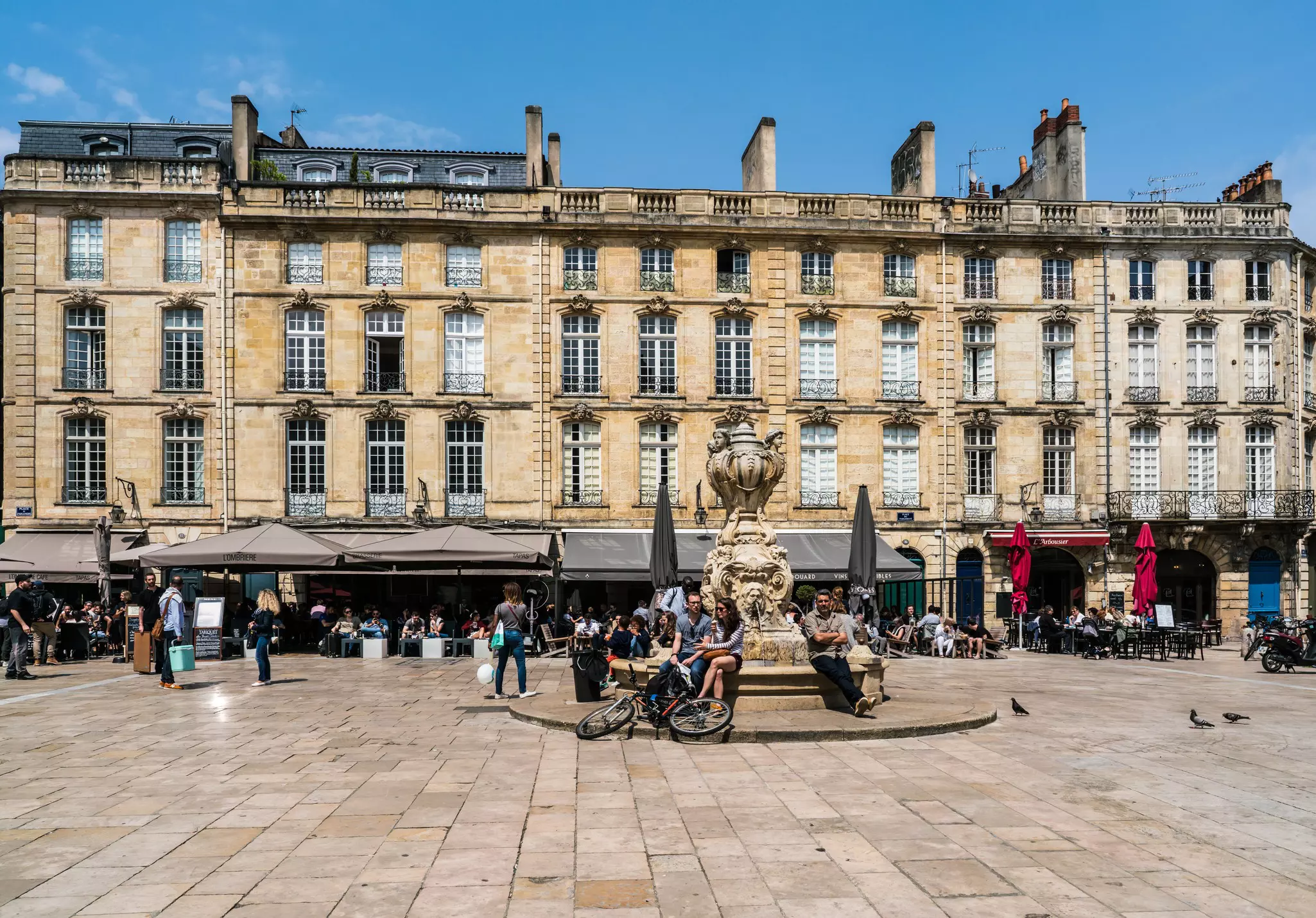 People sit around a fountain and at restaurant terraces in a city with grand architecture.