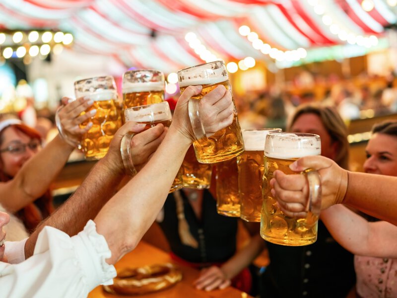 group of freinds in traditional Bavarian attire raising beer steins in a toast at a festive indoor event in an Oktoberfest or dult tent with red and white drapes in the background
