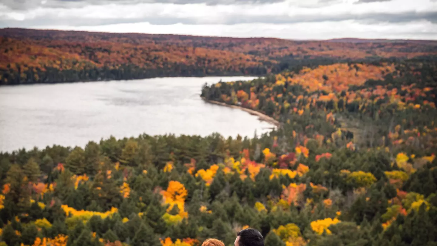 Couple sitting on a rock, overlooking a forest of fall colors and a lake in Algonquin Provincial Park, Canada