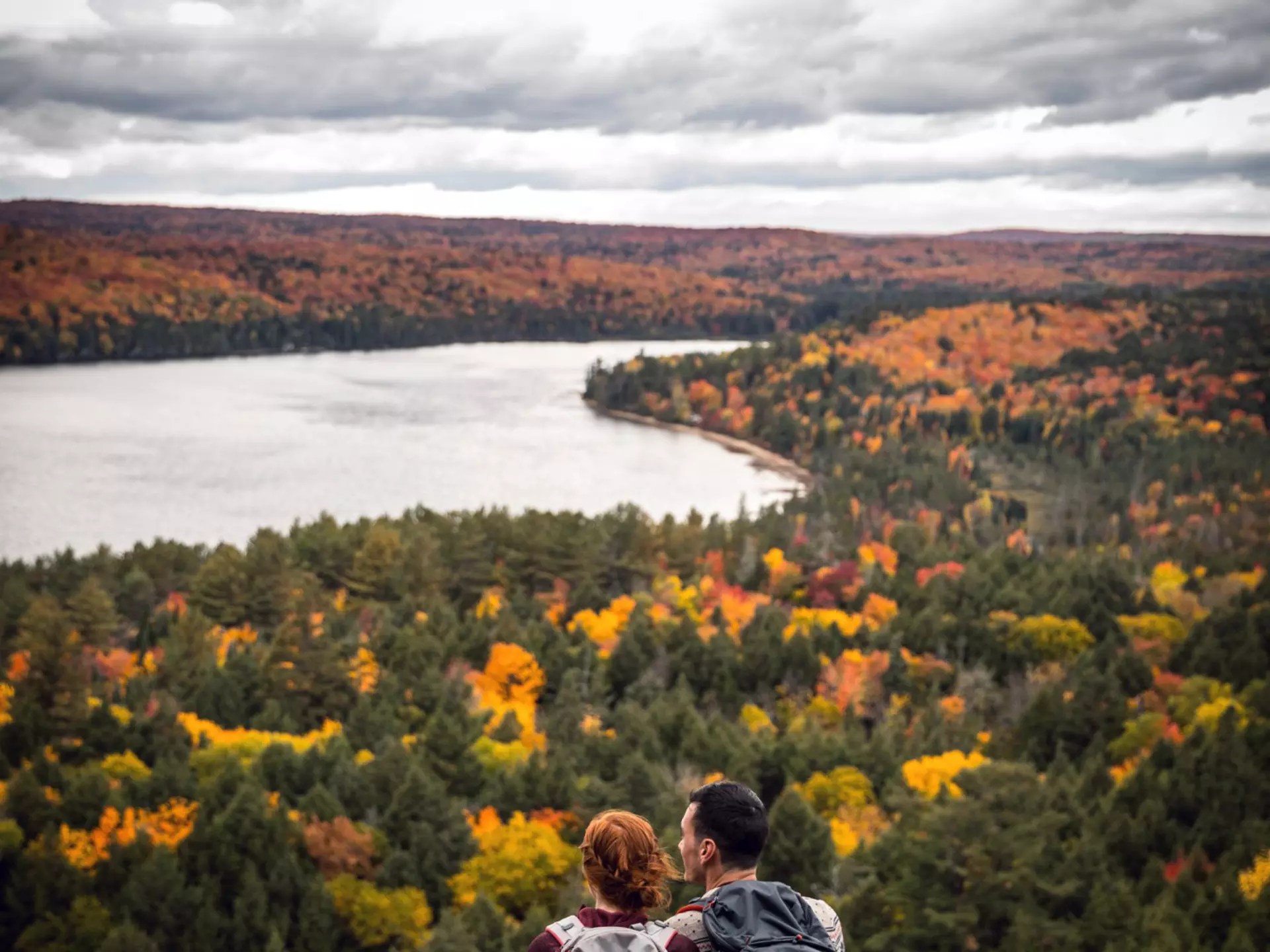 Couple sitting on a rock, overlooking a forest of fall colors and a lake in Algonquin Provincial Park, Canada