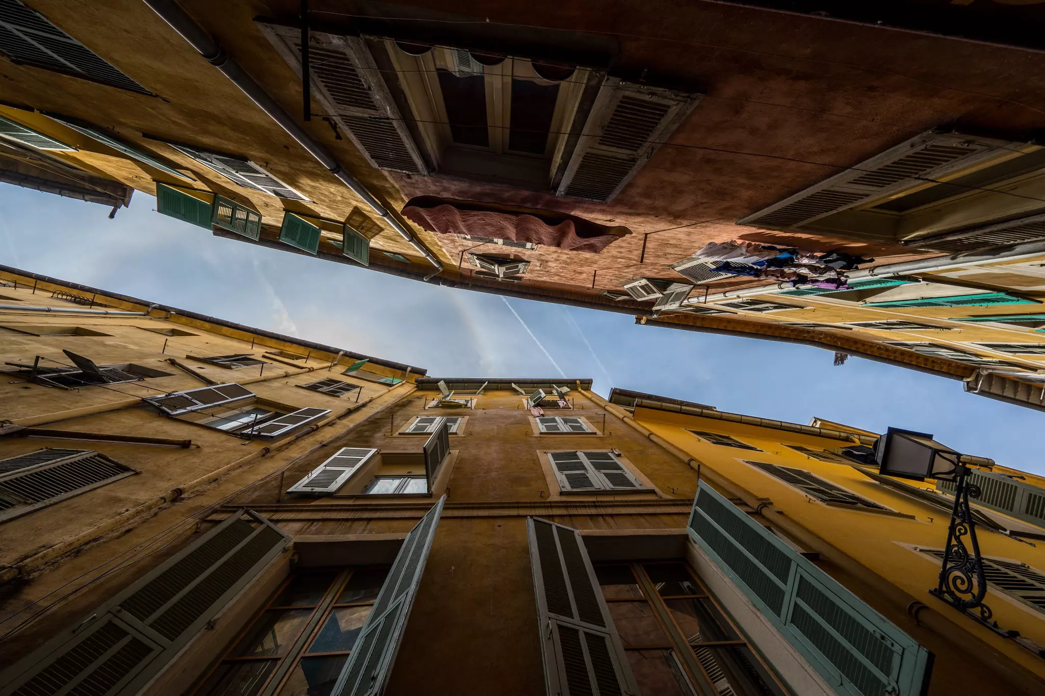 Looking up in a narrow alley, through old buildings with shuttered windows, to a ribbon of blue sky in the old town of Nice, France.