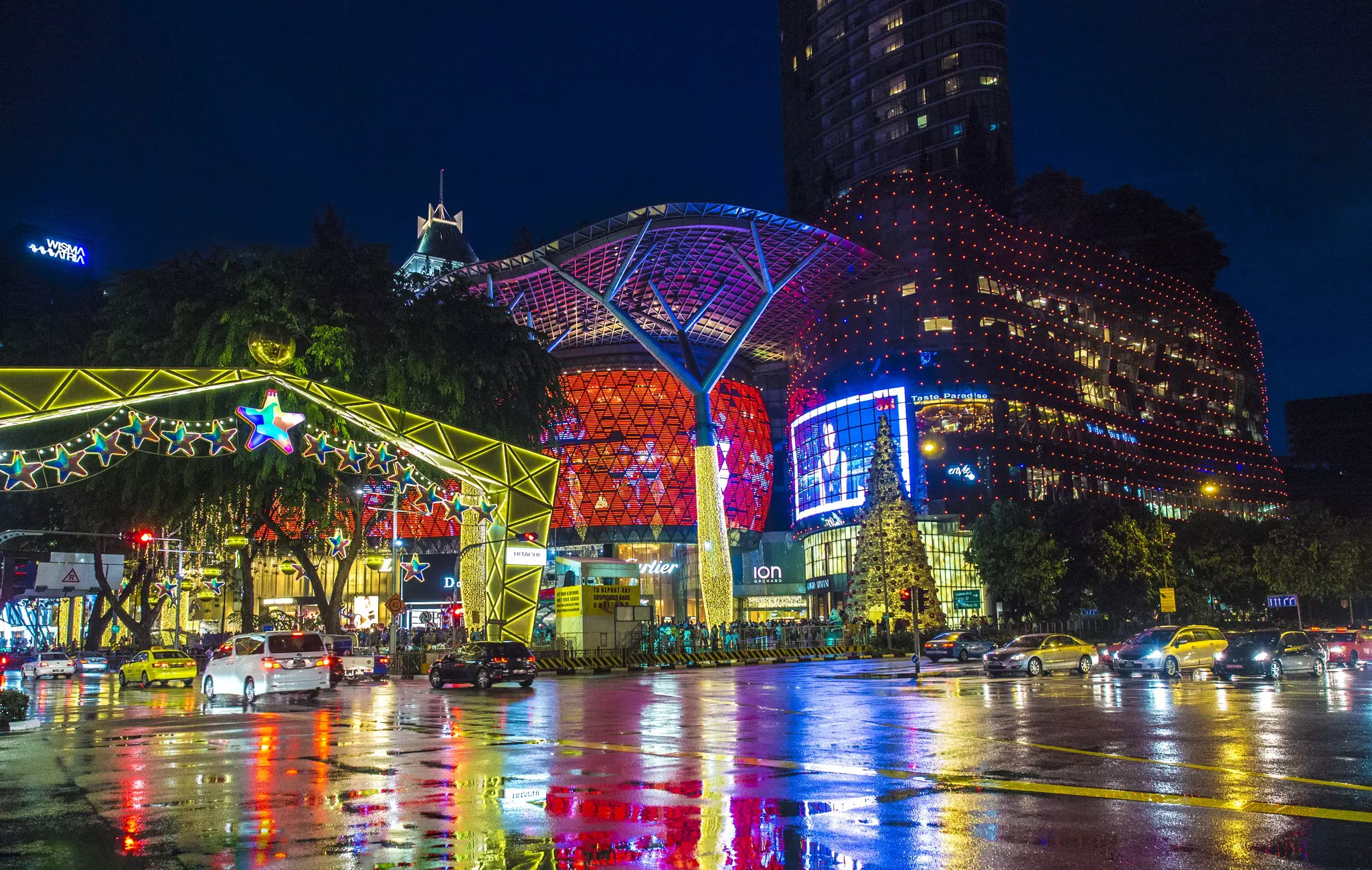 Colorful lights from buildings and roadside decorations reflect on a rainy street at night.
