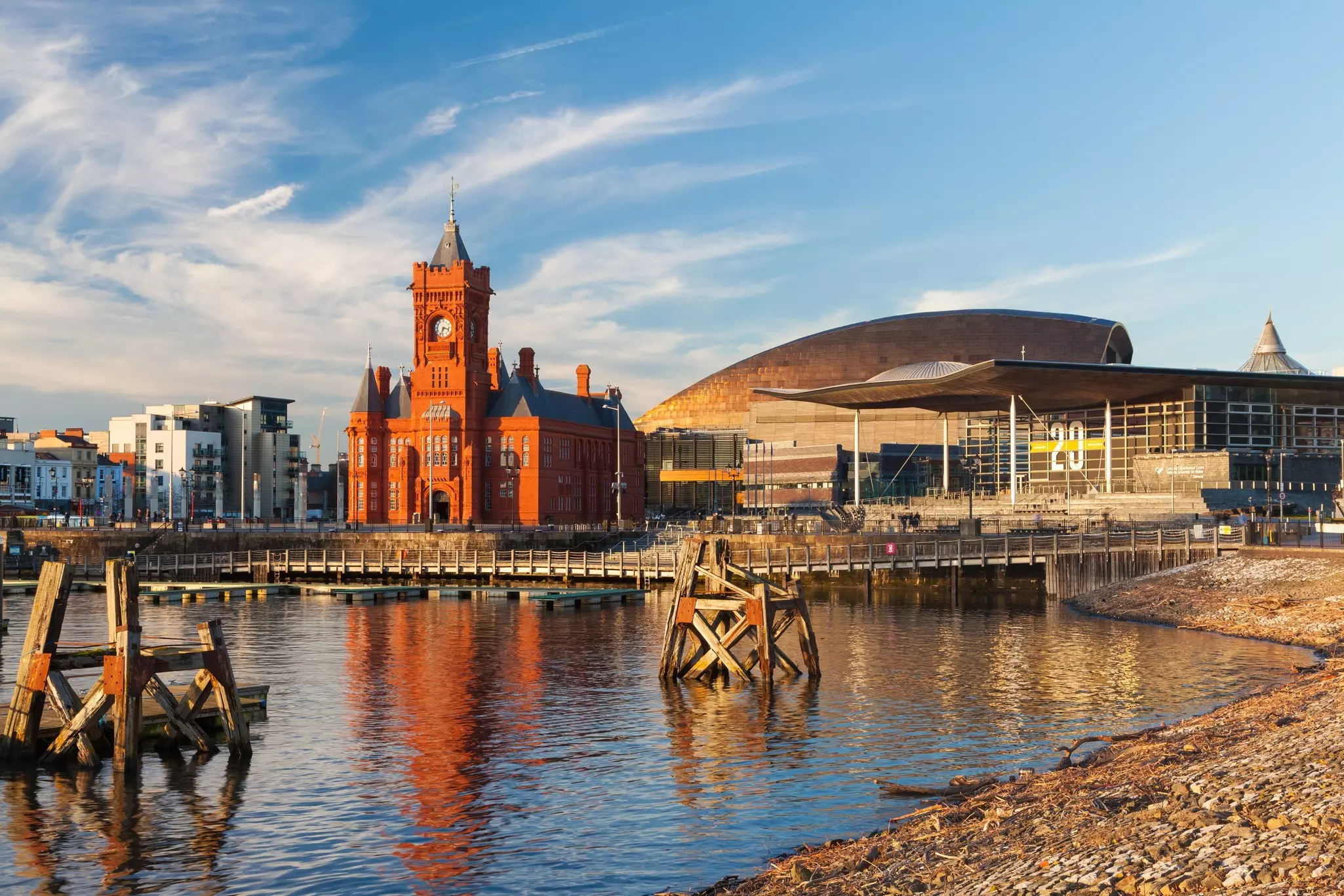 Red castle with clocktower reflected in water surrounded by modern buildings on a sunny day with wisps of clouds in the sky.