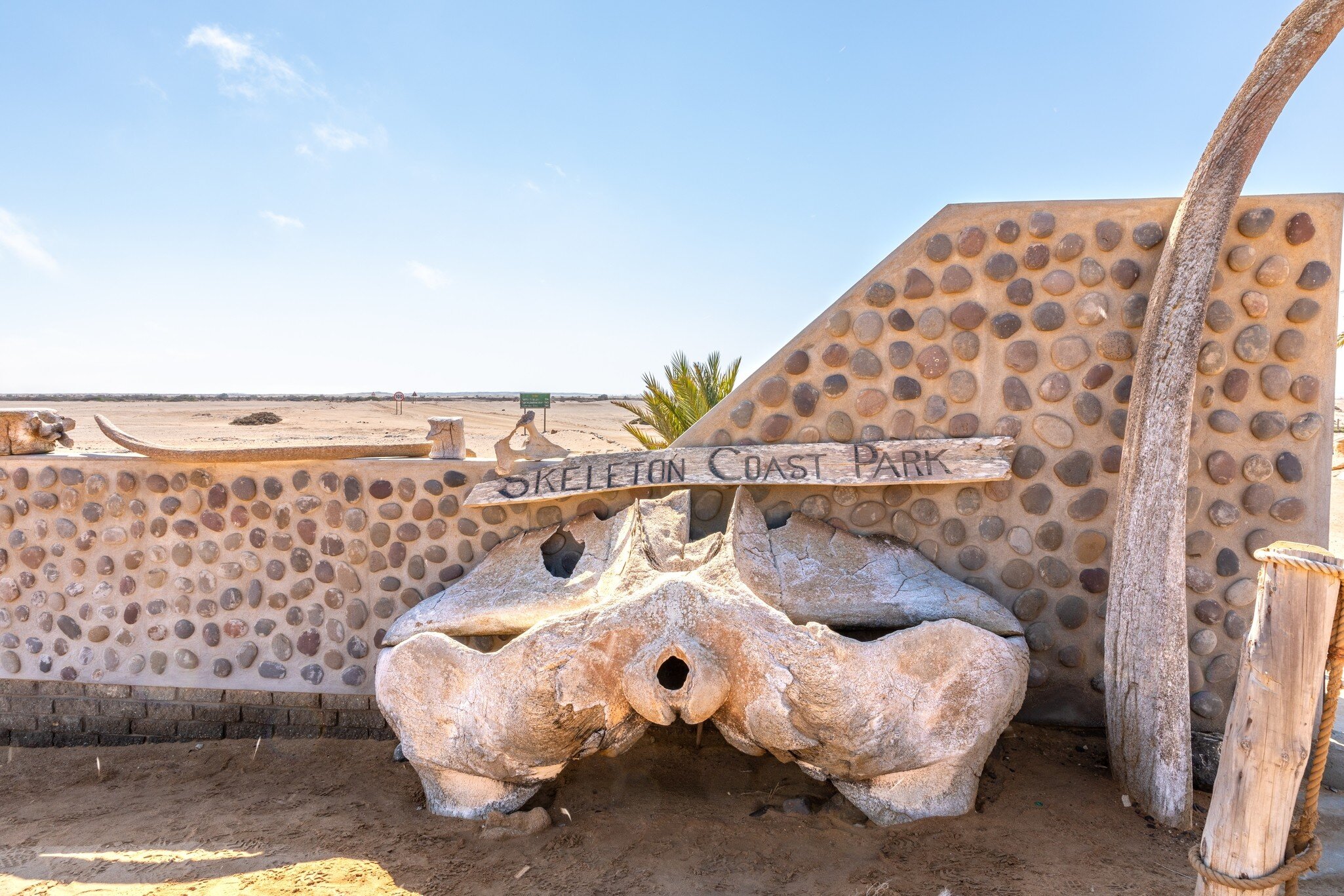 Wooden sign on a pebble-studded wall, with a large bleached bone below and a curved rib beside it in flat desert