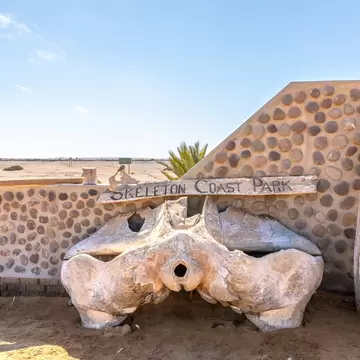 Wooden sign on a pebble-studded wall, with a large bleached bone below and a curved rib beside it in flat desert
