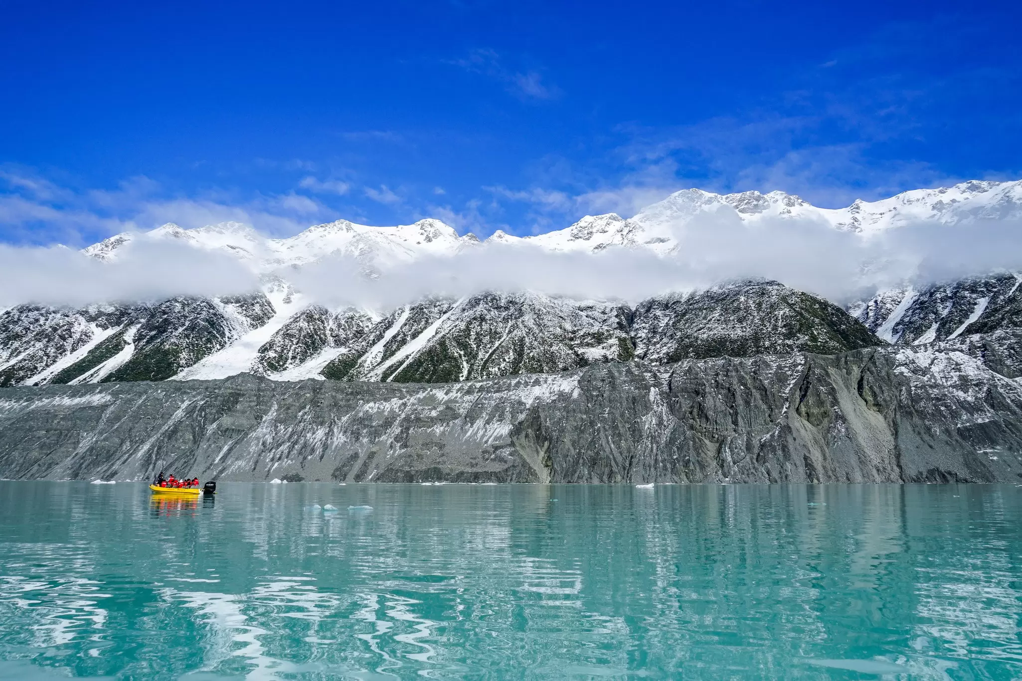 A small yellow boat moving across a blue lake next to a glacier.