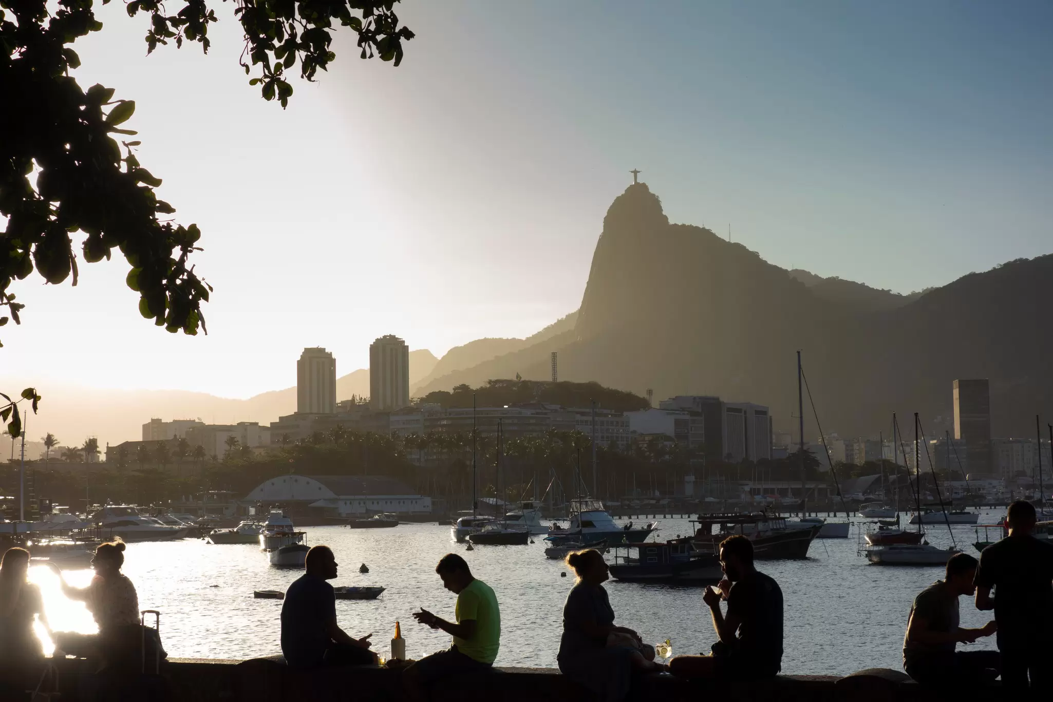 People sitting on a wall by a marina are in silhouette; a mountain with a statue on top is in the background.