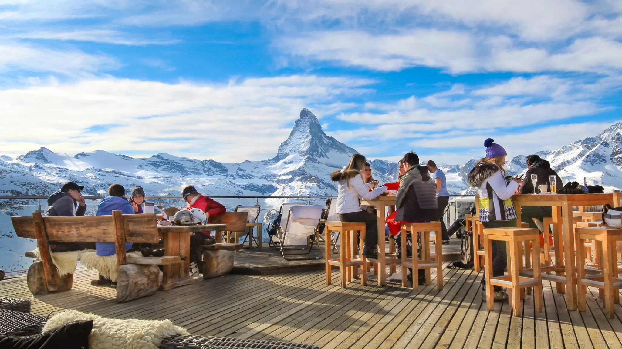 People wearing winter coats sit at wooden tables at an outdoor restaurant overlooking a snowy mountain range in Switzerland.