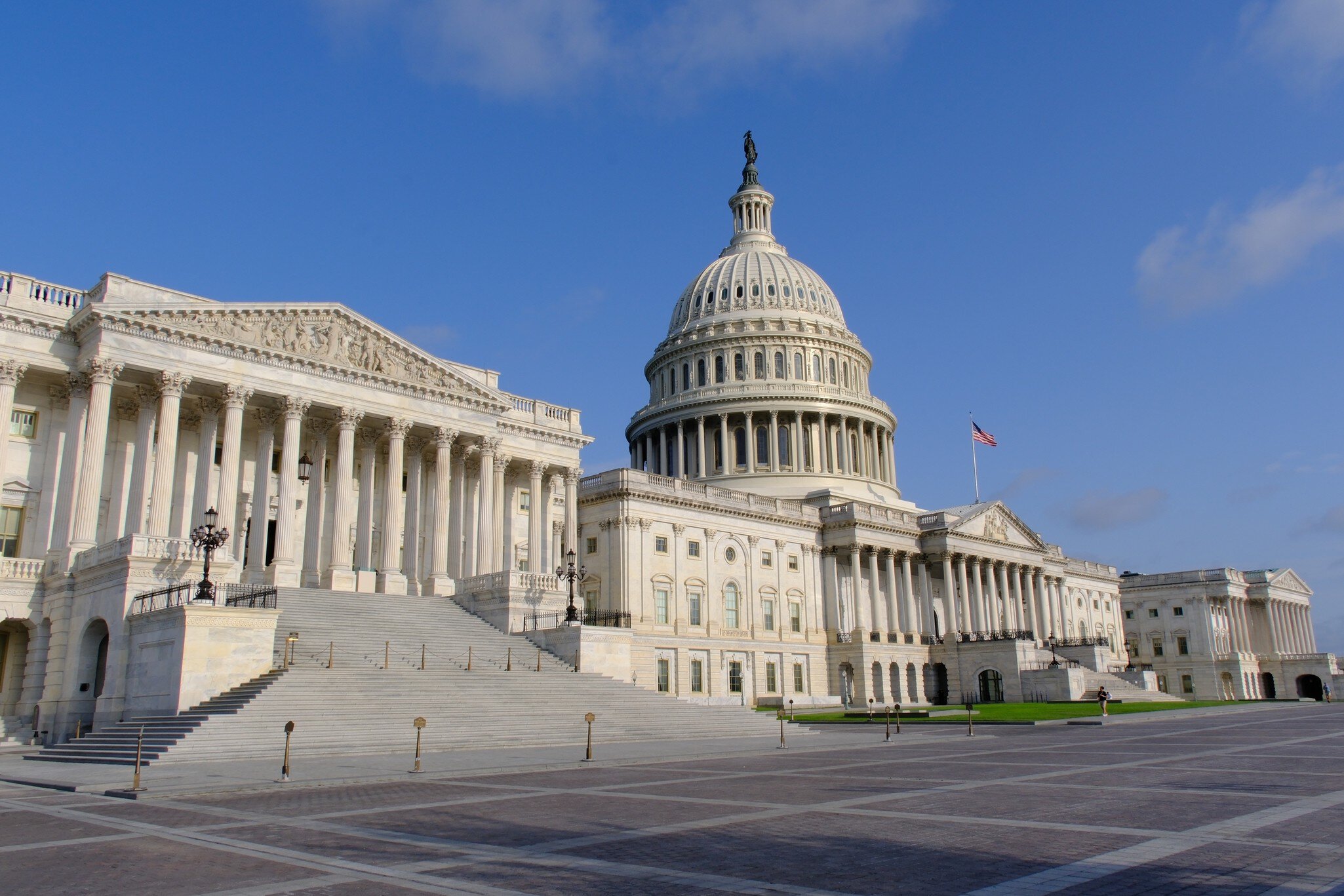 Columned portico and wide marble steps below the statue-topped dome; a US flag over an empty stone plaza.