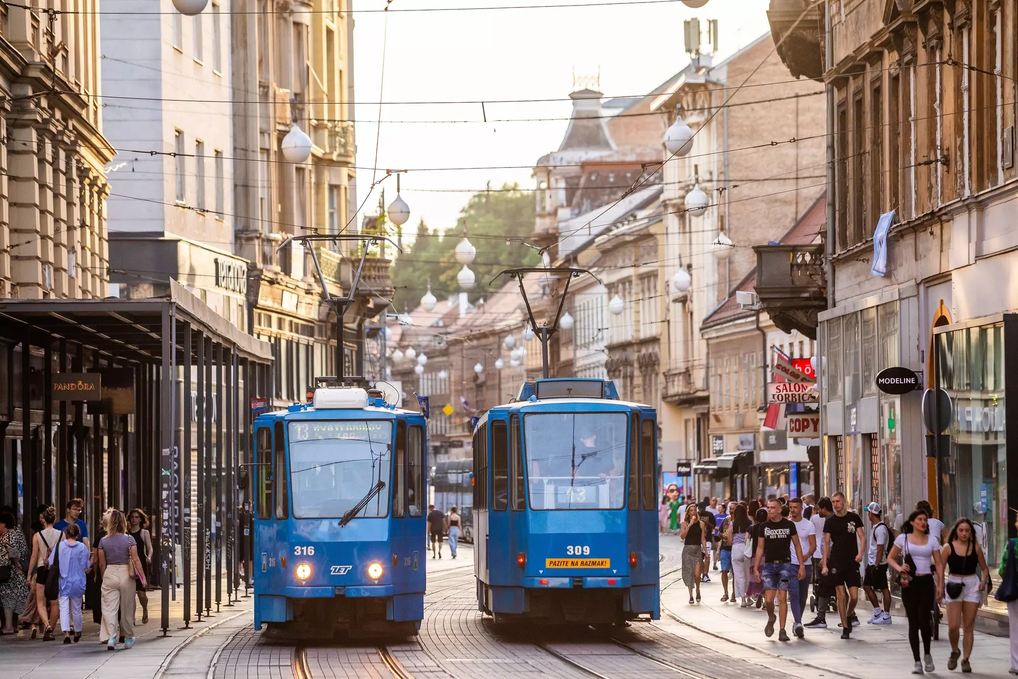 Two blue trams pass one another on a crowded street in a city.