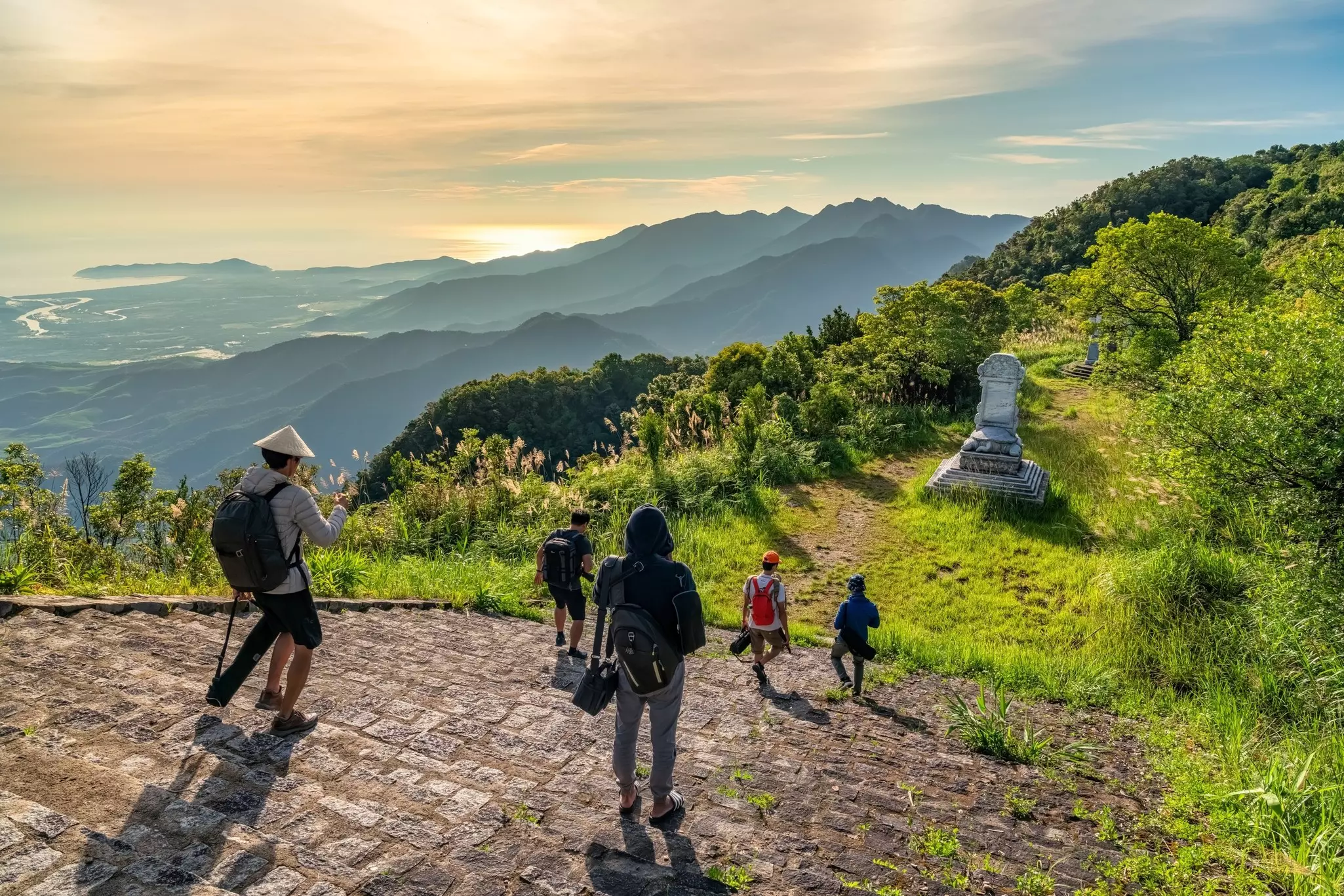 People walk down steps at the top of a hill with views in the distance of the sunrise over the sea.