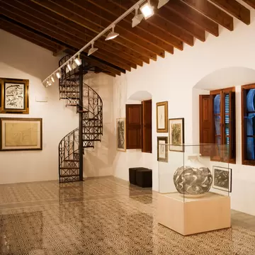White gallery with framed modern artworks, a spiral iron staircase and patterned tiled floor, Sóller, Mallorca
