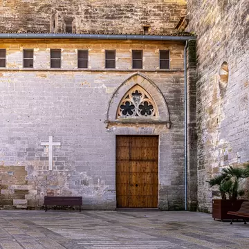 Honey-coloured stone church wall with a pointed arch, trefoil window and studded wooden door, a white cross and benches below