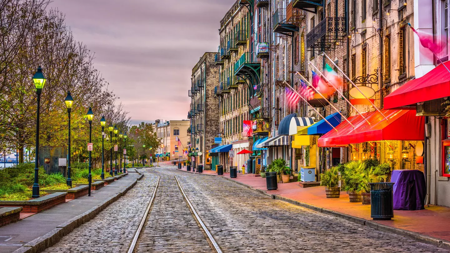 Streetcar tracks over cobblestones pass restaurants and bars on one side and a park with benches on the other under cloudy skies at dusk.