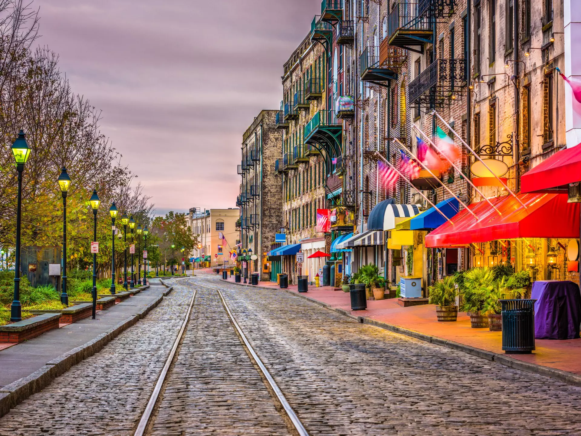 Streetcar tracks over cobblestones pass restaurants and bars on one side and a park with benches on the other under cloudy skies at dusk.