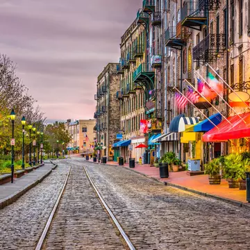 Streetcar tracks over cobblestones pass restaurants and bars on one side and a park with benches on the other under cloudy skies at dusk.