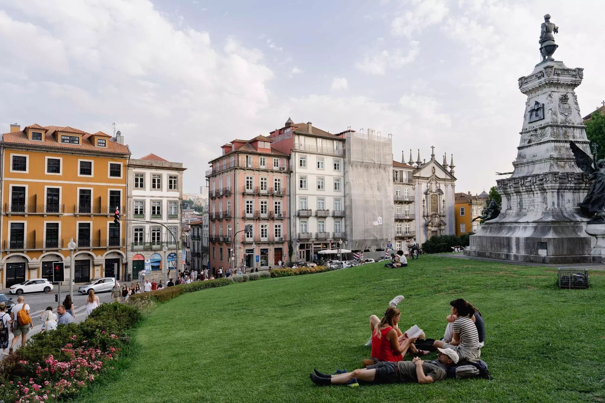 A group of people are sitting and reclining on the green grass of a park in Porto, Portugal; pastel-colored historic buildings are on the opposite side of a street in the background.