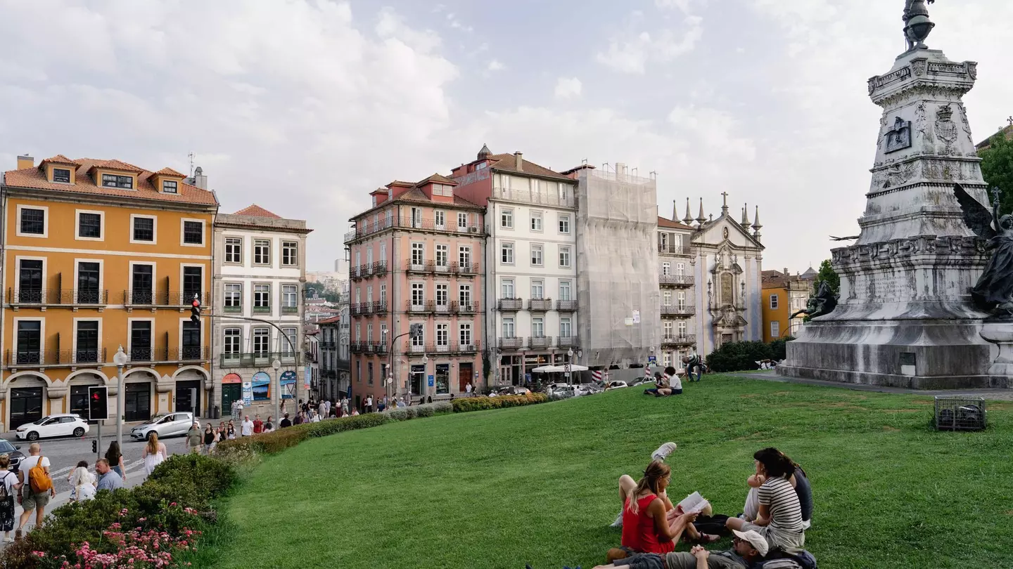 A group of people are sitting and reclining on the green grass of a park in Porto, Portugal; pastel-colored historic buildings are on the opposite side of a street in the background.