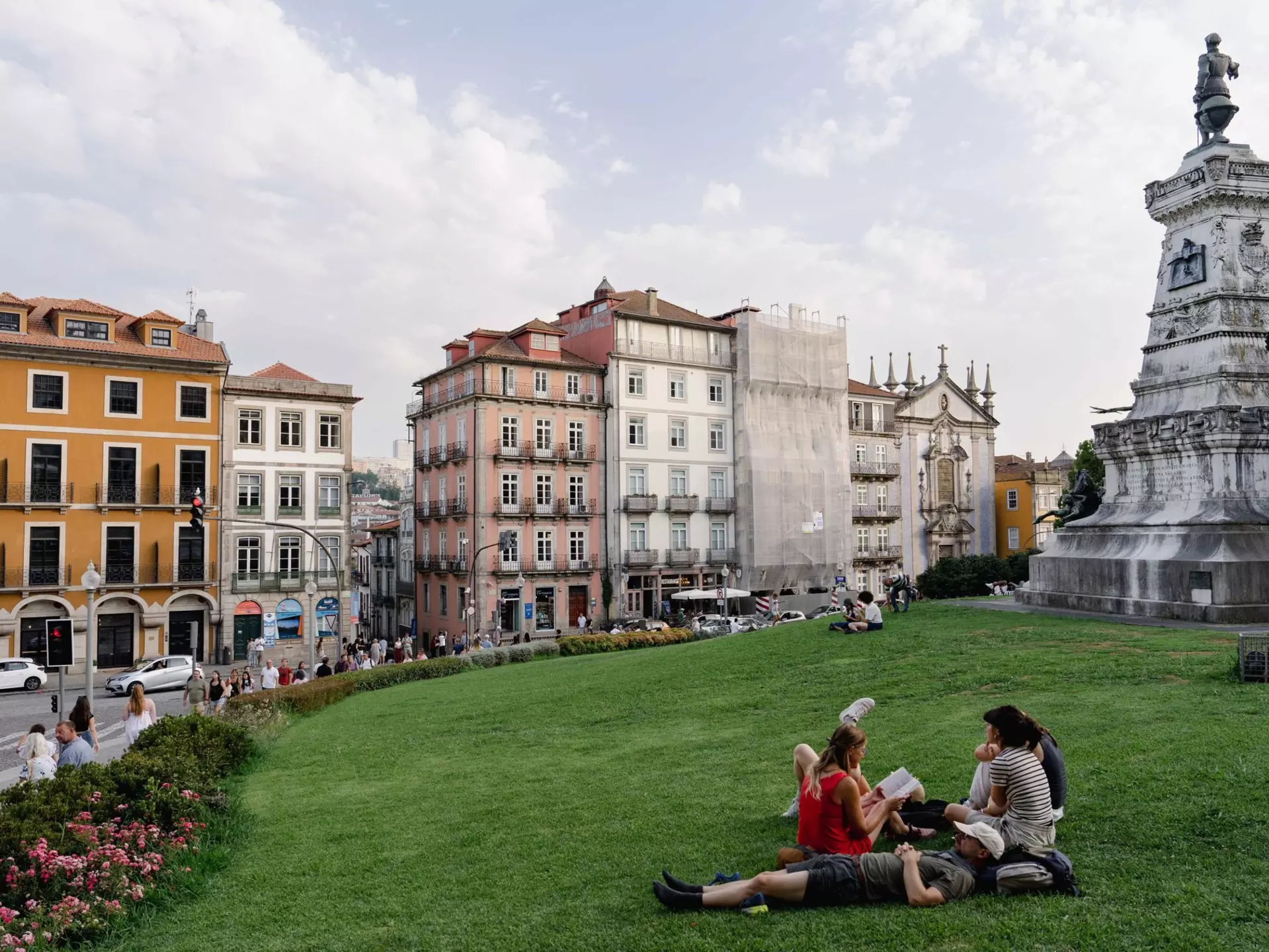 A group of people are sitting and reclining on the green grass of a park in Porto, Portugal; pastel-colored historic buildings are on the opposite side of a street in the background.