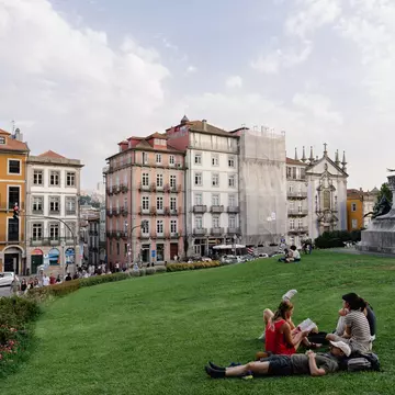 A group of people are sitting and reclining on the green grass of a park in Porto, Portugal; pastel-colored historic buildings are on the opposite side of a street in the background.