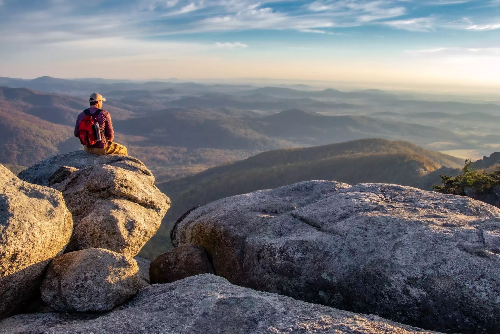 Man on boulders overlooking forested mountains at sunrise.