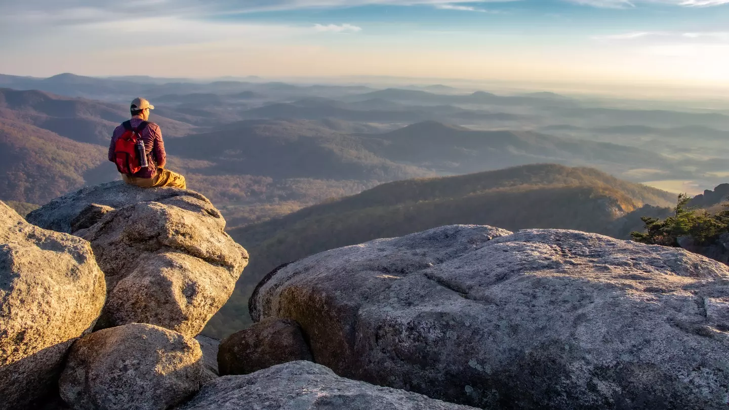Man on boulders overlooking forested mountains at sunrise.