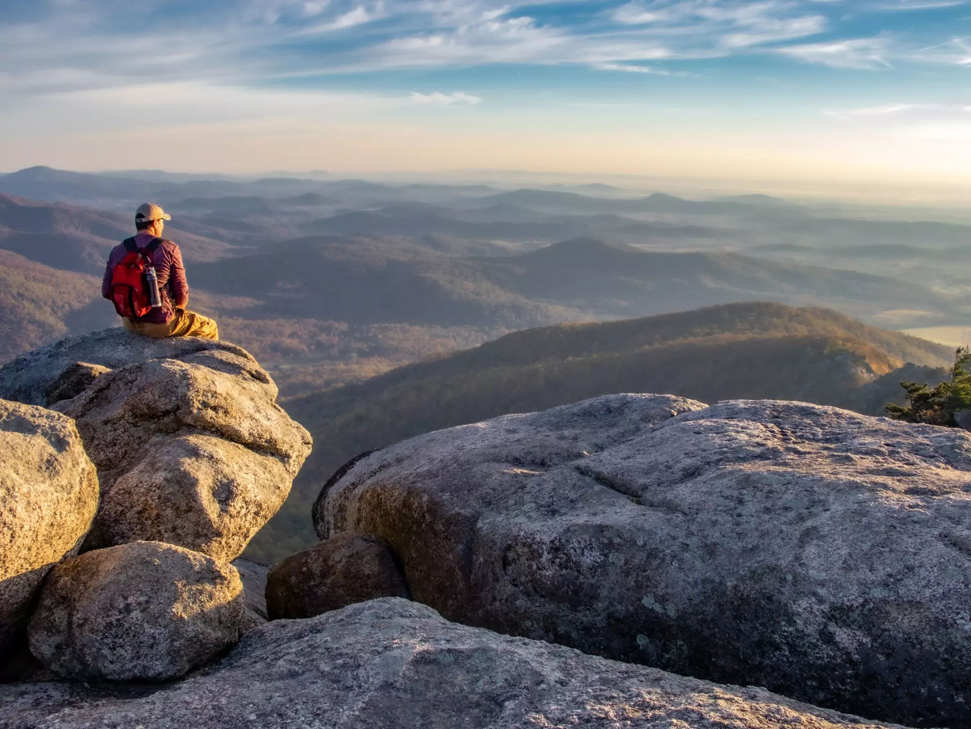Man on boulders overlooking forested mountains at sunrise.