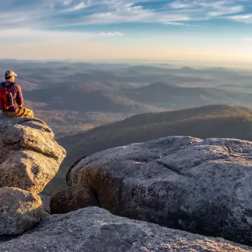 Man on boulders overlooking forested mountains at sunrise.