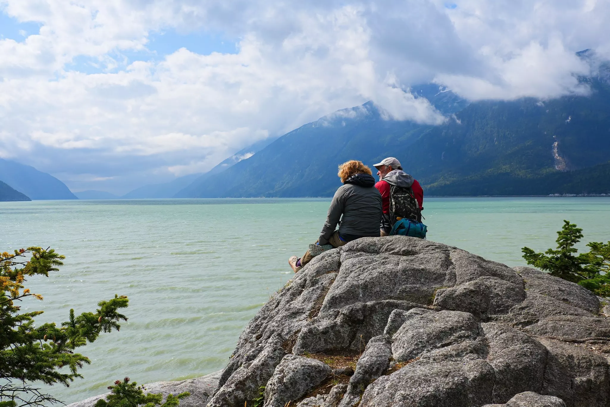 Two people in jackets sit on a boulder by a turquoise lake; mountains in clouds are on the far shore.