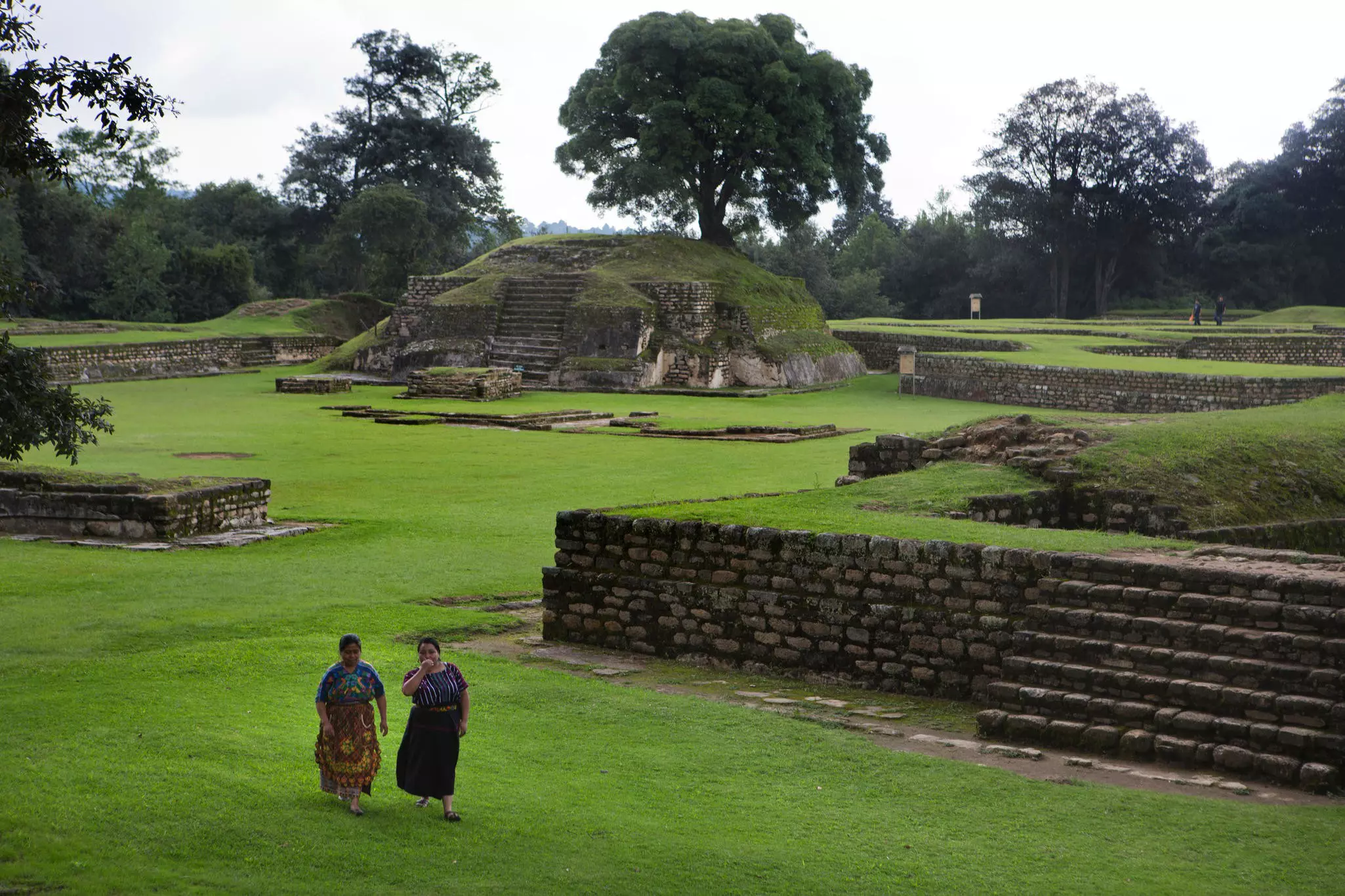 Two woman walk through an archeological park with stone platforms and tidy lawns.