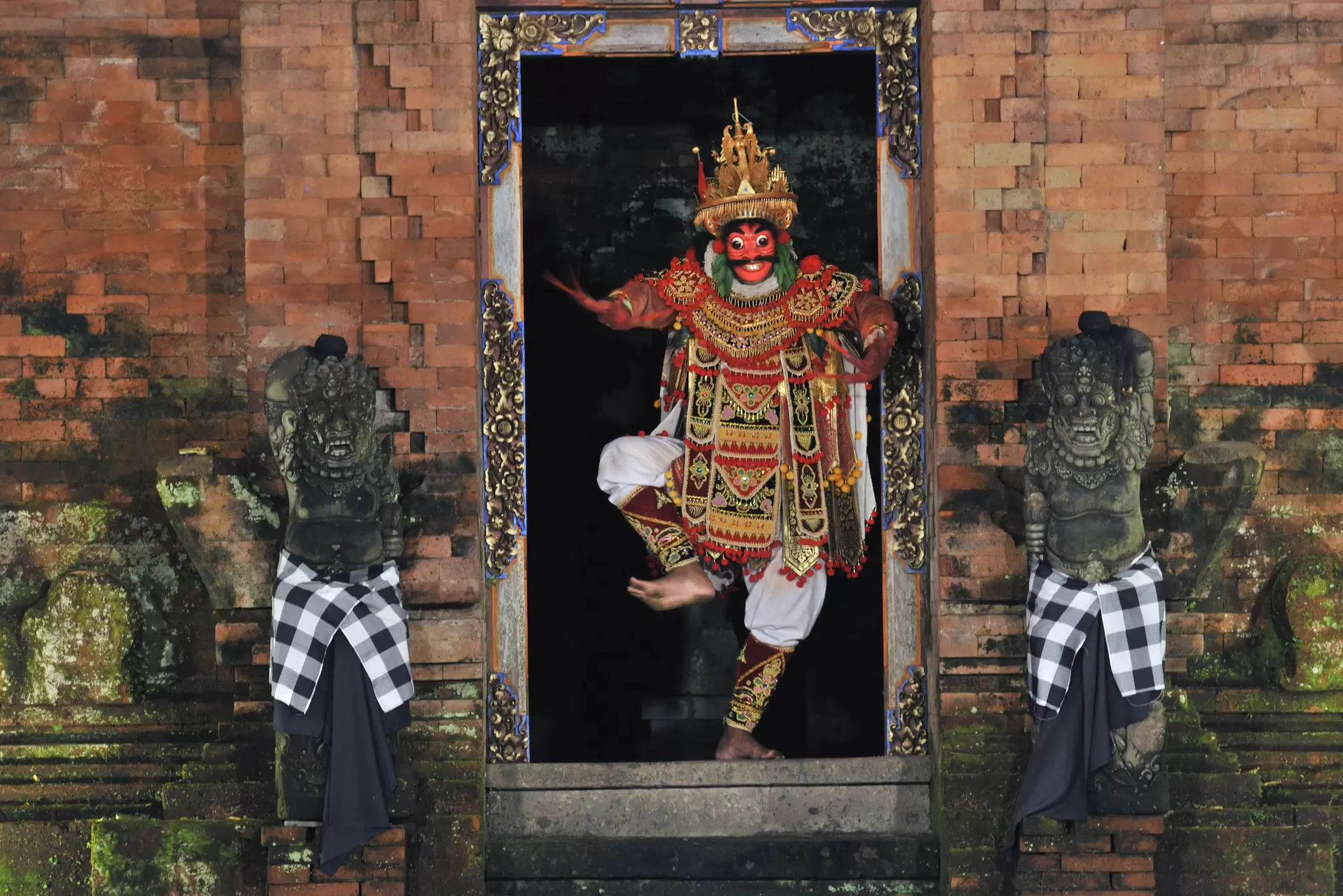 A Balinese performer wearing red facepaint and a colored costume dances between two statues.
