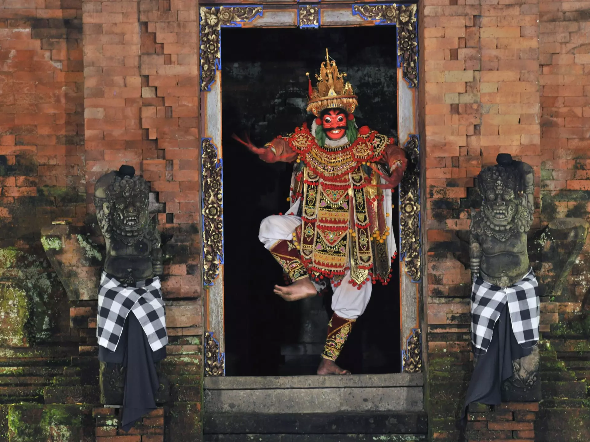 A Balinese performer wearing red facepaint and a colored costume dances between two statues.