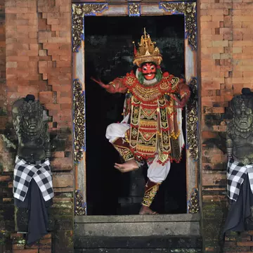 A Balinese performer wearing red facepaint and a colored costume dances between two statues.