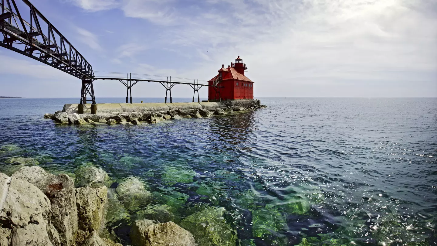 A red, squat lighthouse building at the edge of pier leading into a lake
