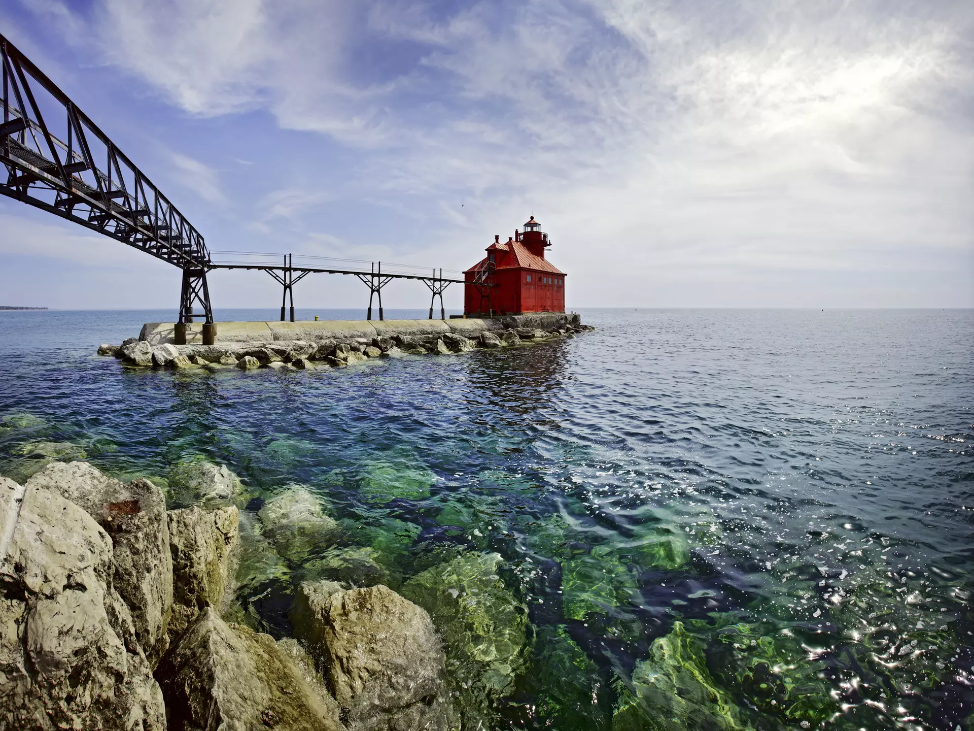 A red, squat lighthouse building at the edge of pier leading into a lake