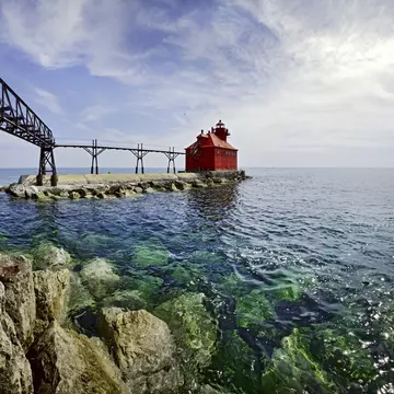A red, squat lighthouse building at the edge of pier leading into a lake