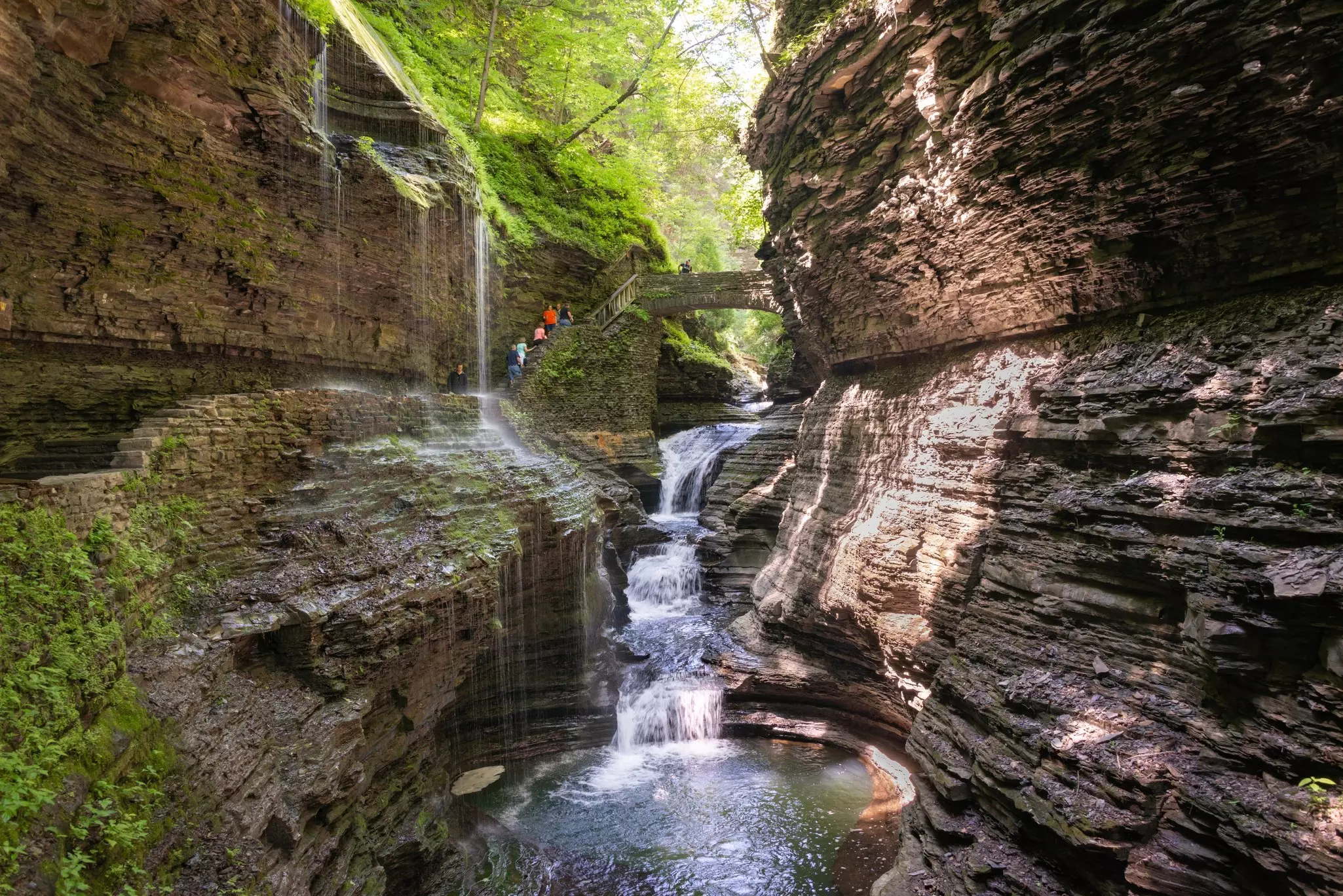 Hikers in New York climb toward a stone bridge over a narrow gorge with a waterfall at the center.