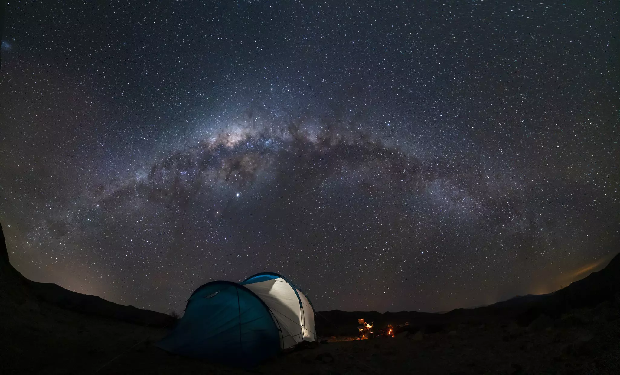 A tent, a campfire and a dark sky lit up with stars with the sweep of the Milky Way