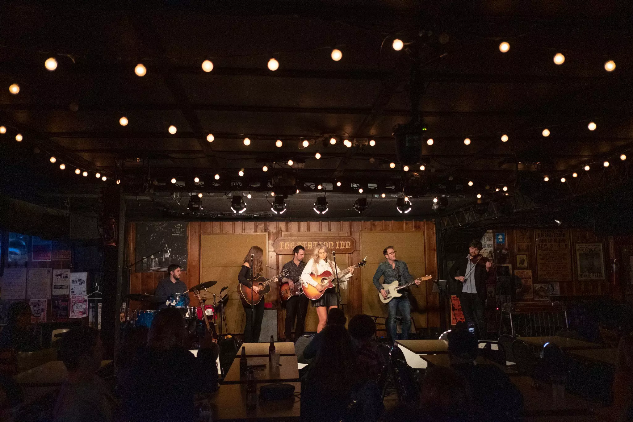 A female performer fronts a band on the stage of a small indoor music venue.