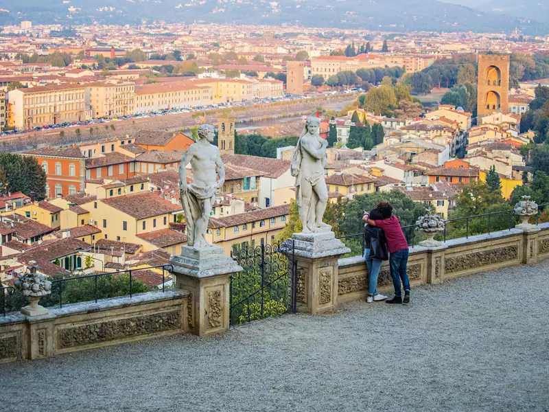 Two people lean against the wall of an overlook. A wide view of a city with red-roofed buildings and church towers is visible from the terrace.