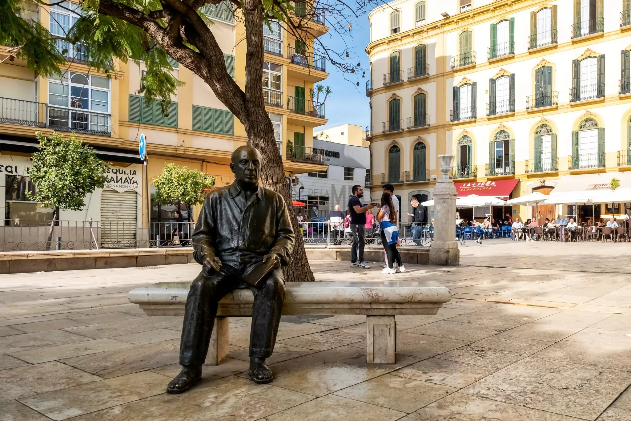 A bronze life-size sculpture of a man is set on a bench in a city square.
