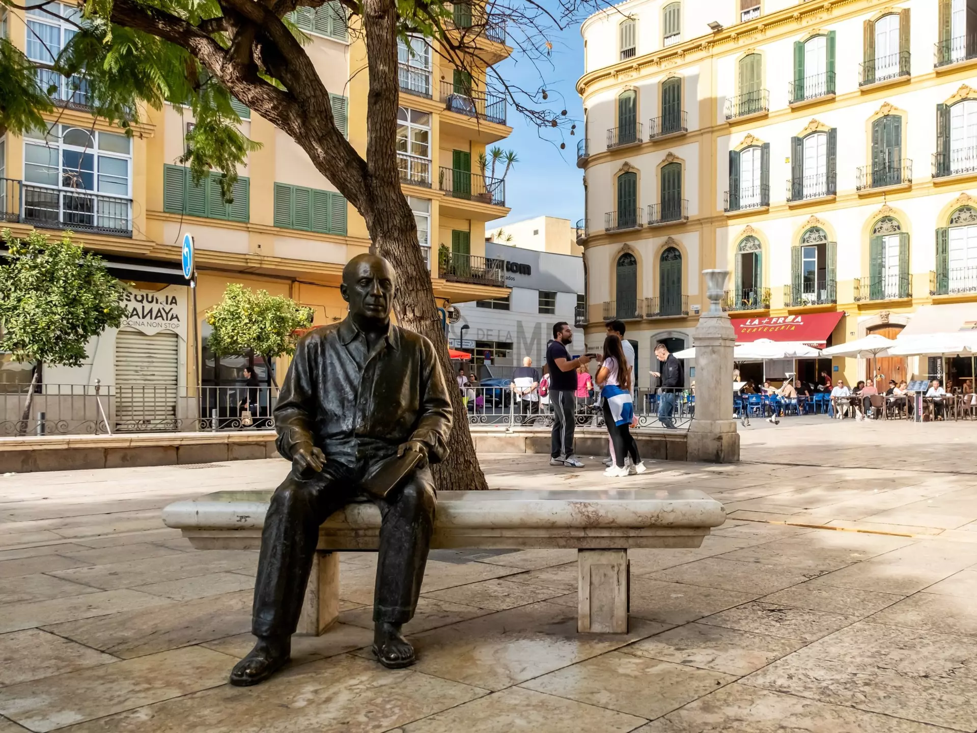 A bronze life-size sculpture of a man is set on a bench in a city square.