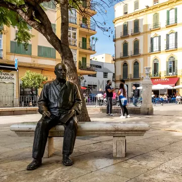 A bronze life-size sculpture of a man is set on a bench in a city square.