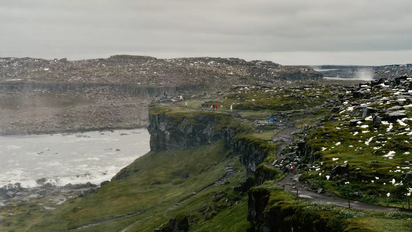 People in the far distance on a path atop a grass-covered rocky hill with water and other rocky hills beyond on a misty, overcast day.